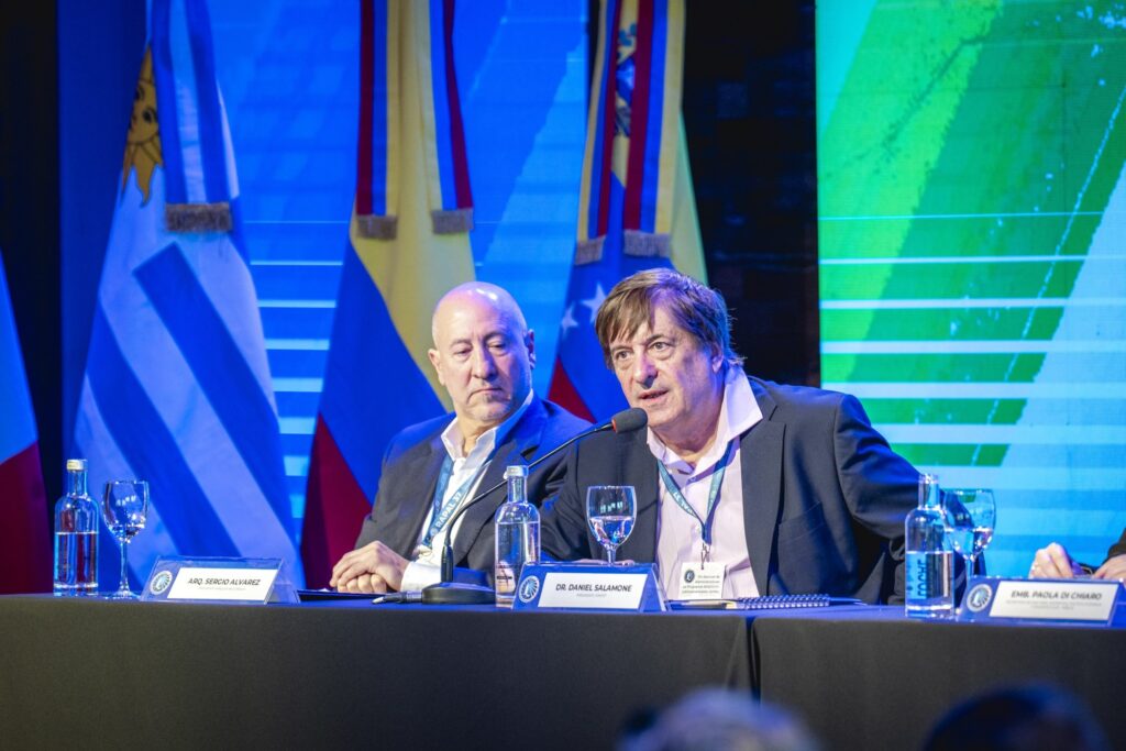 Dos autoridades en la mesa de apertura de la sesión plenaria: una habla al micrófono y la otra la escucha, frente a las banderas de los países