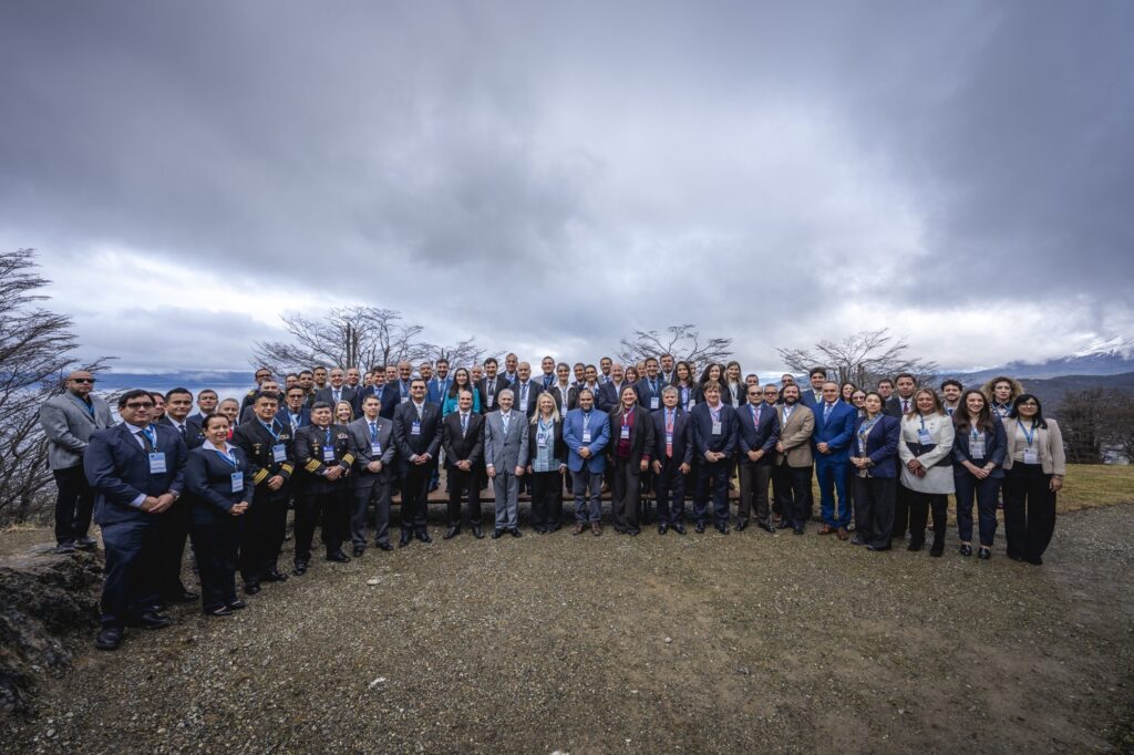 Foto oficial de los participantes de la XXXVII RAPAL, reunidos al aire libre bajo un cielo nublado, con la bahía y un monte nevado detrás