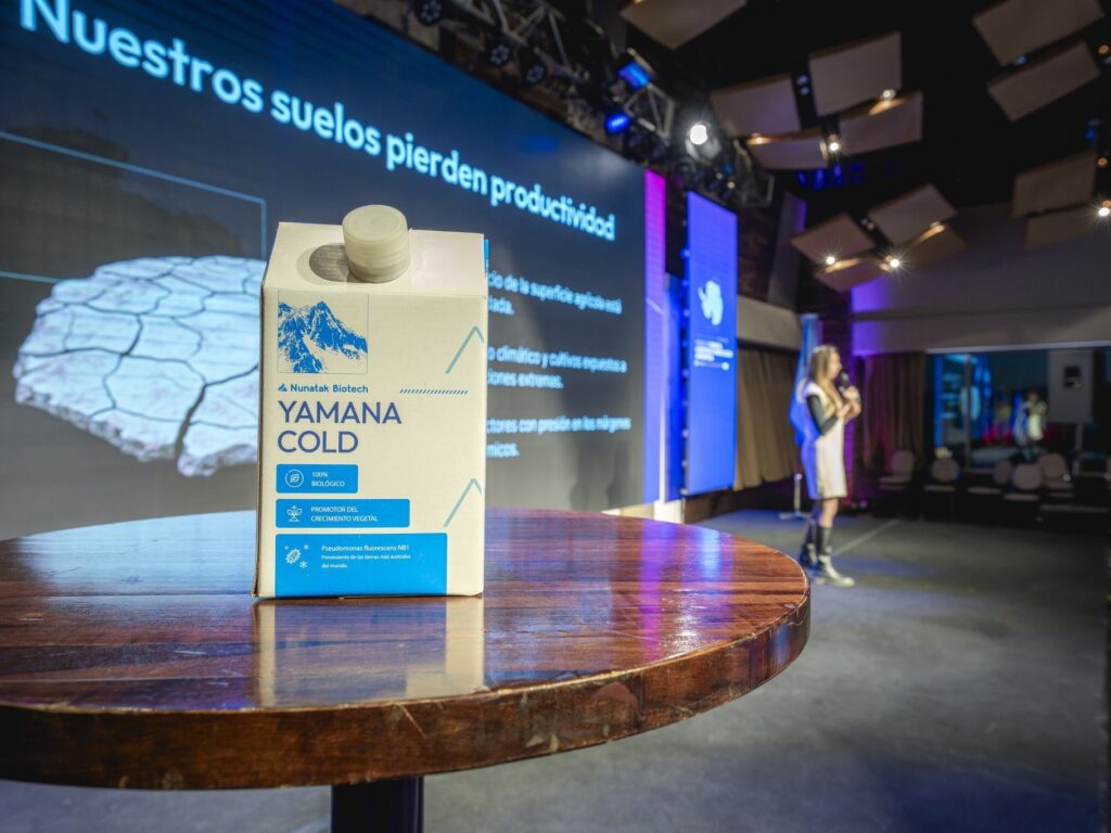 Envase de un bioinsumo de Nunatak sobre una mesa del escenario, con una presentadora y la pantalla de fondo
