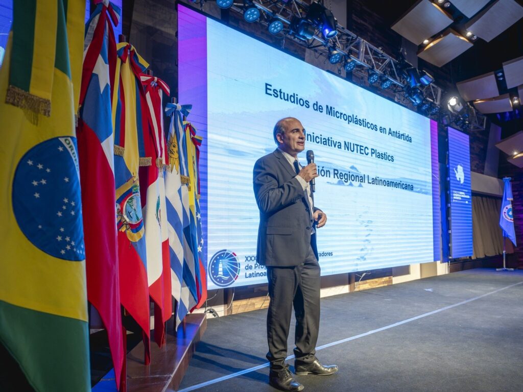 Expositor de NUTEC Plastics en el escenario junto a las banderas de los países, frente a una pantalla sobre microplásticos en la Antártida