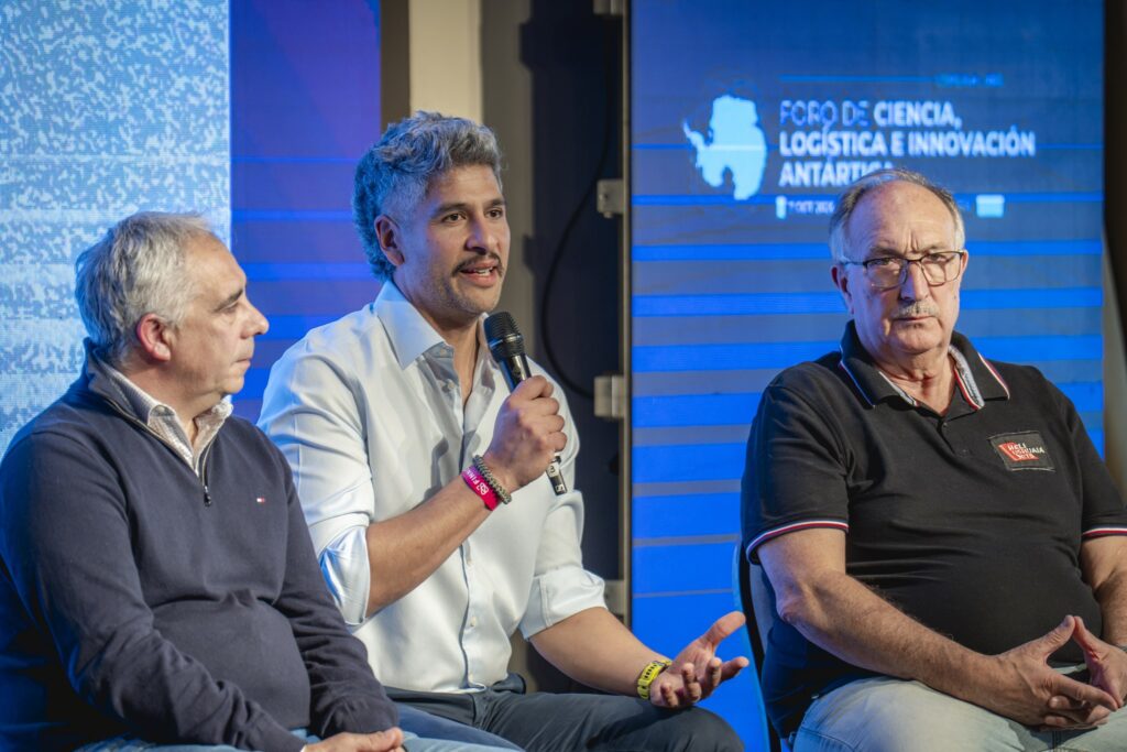 Tres panelistas en el escenario del Foro, uno de ellos hablando por micrófono, con el logo del Foro detrás