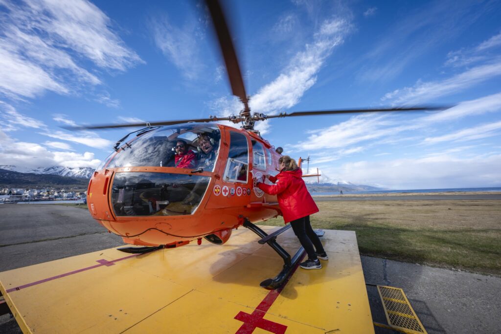 Helicóptero naranja con las hélices en movimiento sobre la plataforma del helipuerto de Ushuaia, con dos personas a bordo y otra junto a la puerta